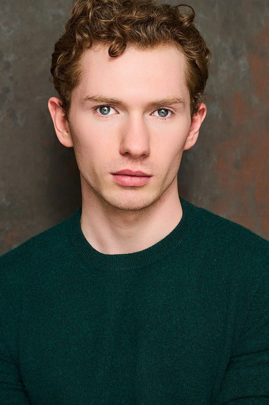 Close-up portrait of a young man with curly light brown hair, blue eyes, fair skin, wearing a dark green sweater, against a painted wall background.