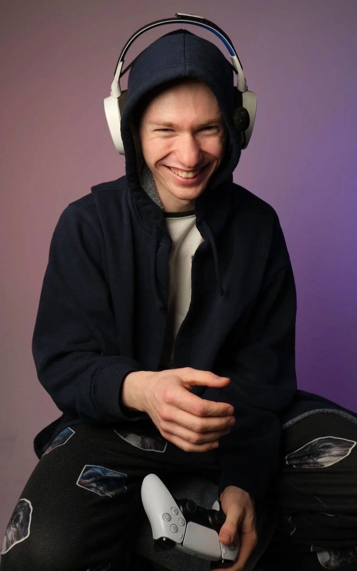 A young man smiling while sitting against a purple gradient background. He's wearing a black hoodie, a dark jacket, and holding a video game controller in his right hand.