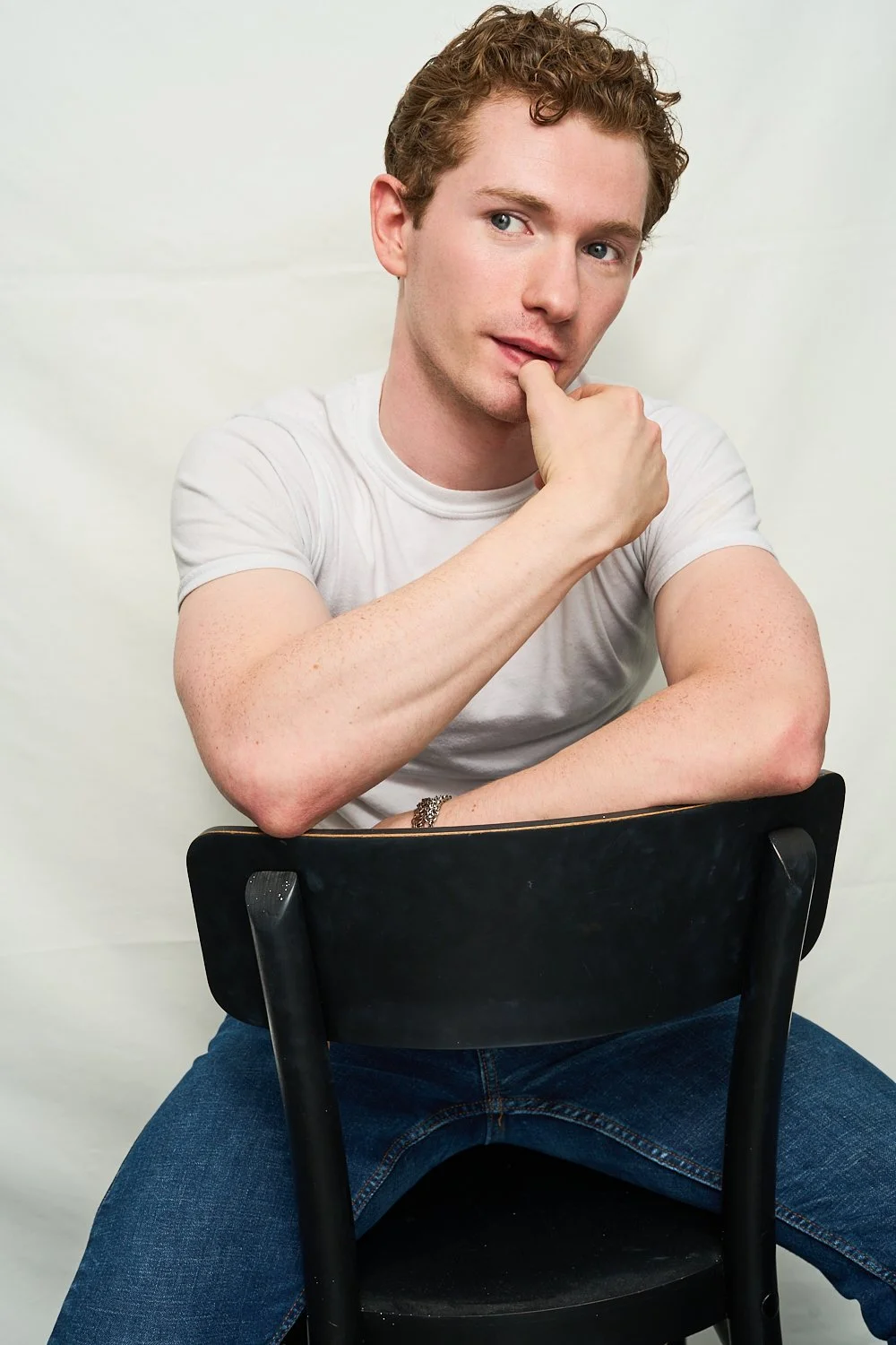 Young man sitting backwards on a black chair, wearing a white T-shirt and blue jeans, with his right arm resting on the chair's backrest, looking thoughtfully into the distance with his chin resting on his left hand.