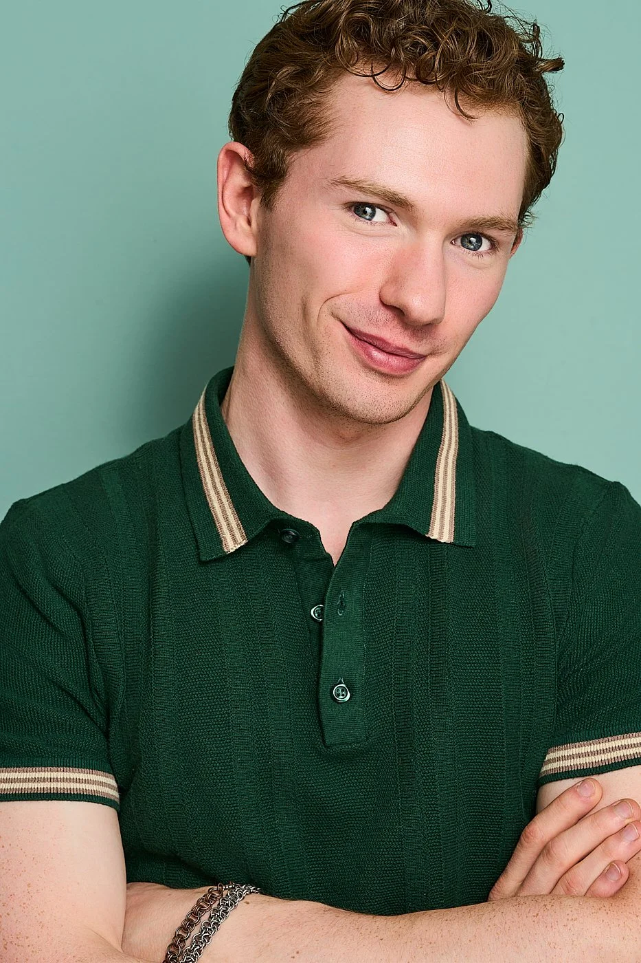 A young man with curly brown hair, blue eyes, and fair skin is smiling slightly, looking directly at the camera. He's wearing a dark green polo shirt with beige and black striped accents on the collar and sleeves, and has a chain bracelet on his wris
