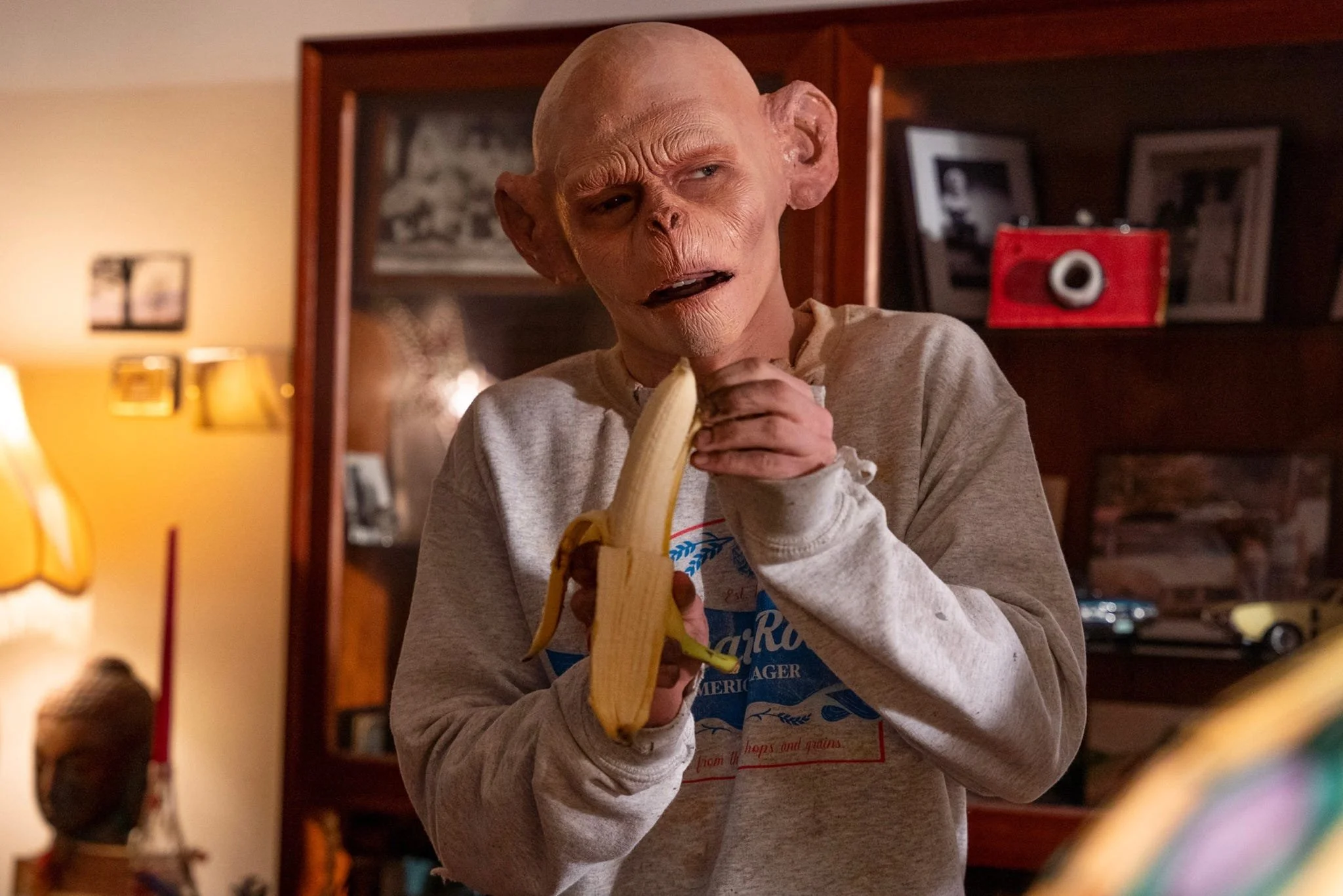 A person with a deformed face and bald head holding a banana. They are in a room with wooden shelves and framed pictures in the background, wearing a gray sweatshirt.