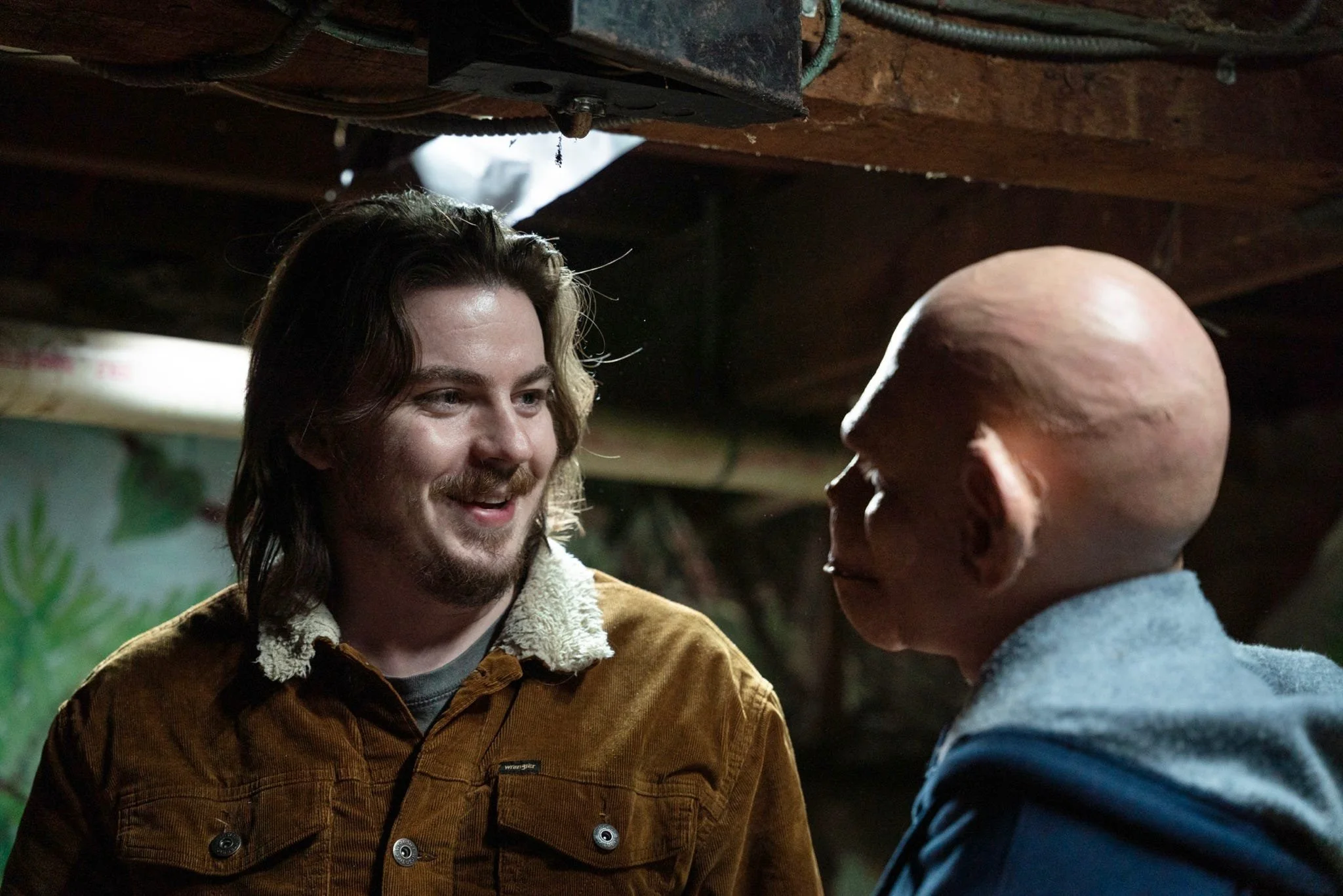 Two men are smiling and facing each other in a dimly lit setting, possibly indoors with wooden ceiling beams visible.
