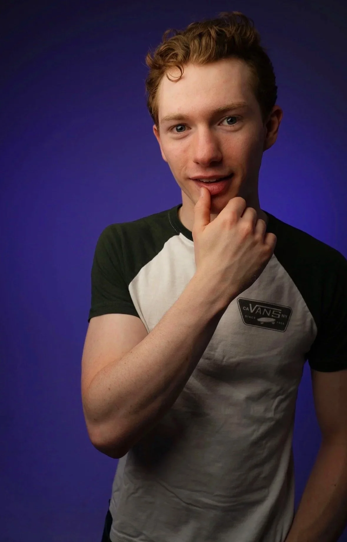 Young man with curly red hair wearing a gray and black Vans t-shirt, posing with his finger on his lips against a blue background.