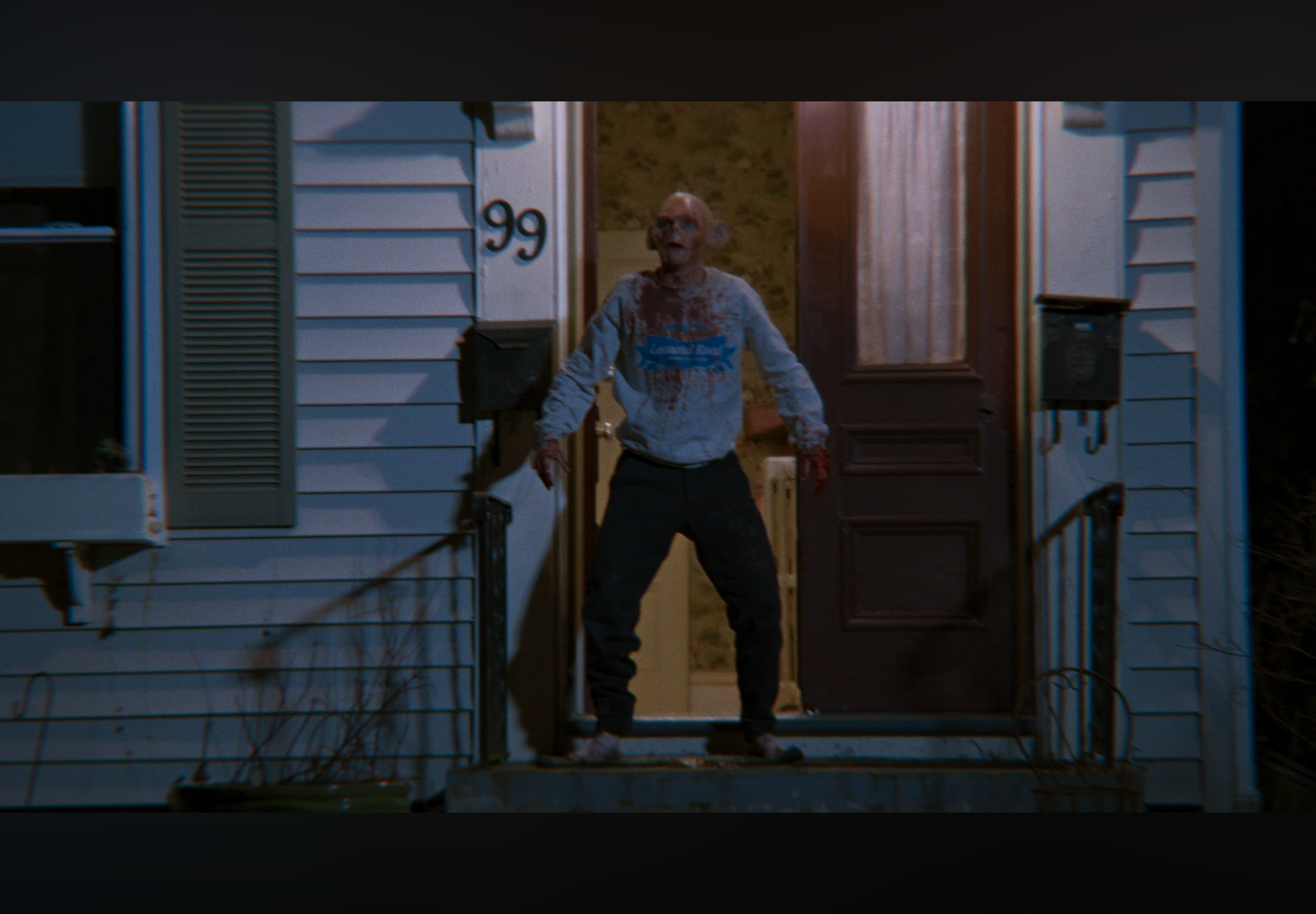 A person standing on a porch at night, wearing a blood-splattered sweatshirt, with a bloodstained face and hands, looking surprised or scared.