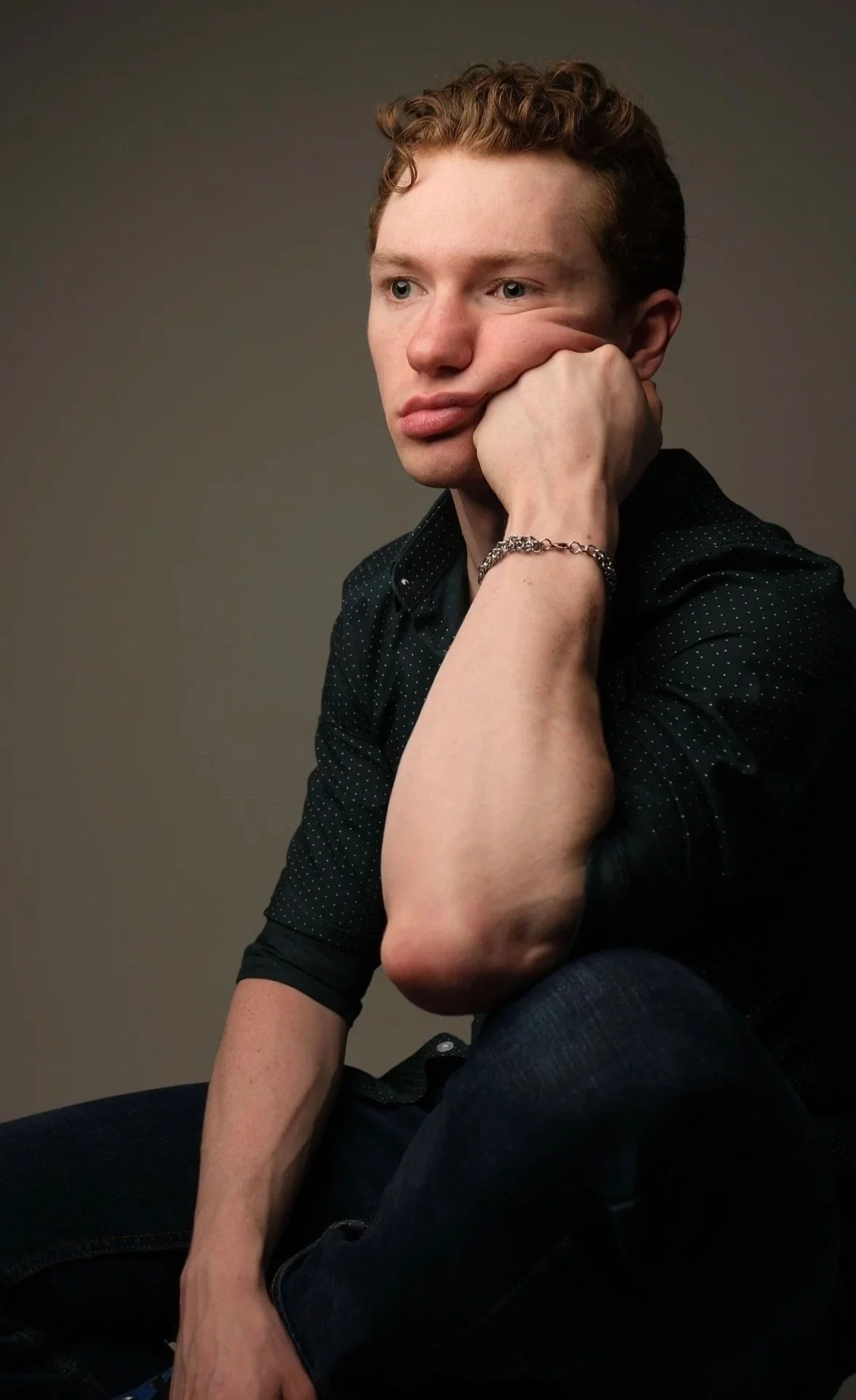 Portrait of a young man with light skin and short curly hair, sitting with his hand on his face, wearing a dark polka-dot shirt and a silver chain bracelet, against a plain dark background.