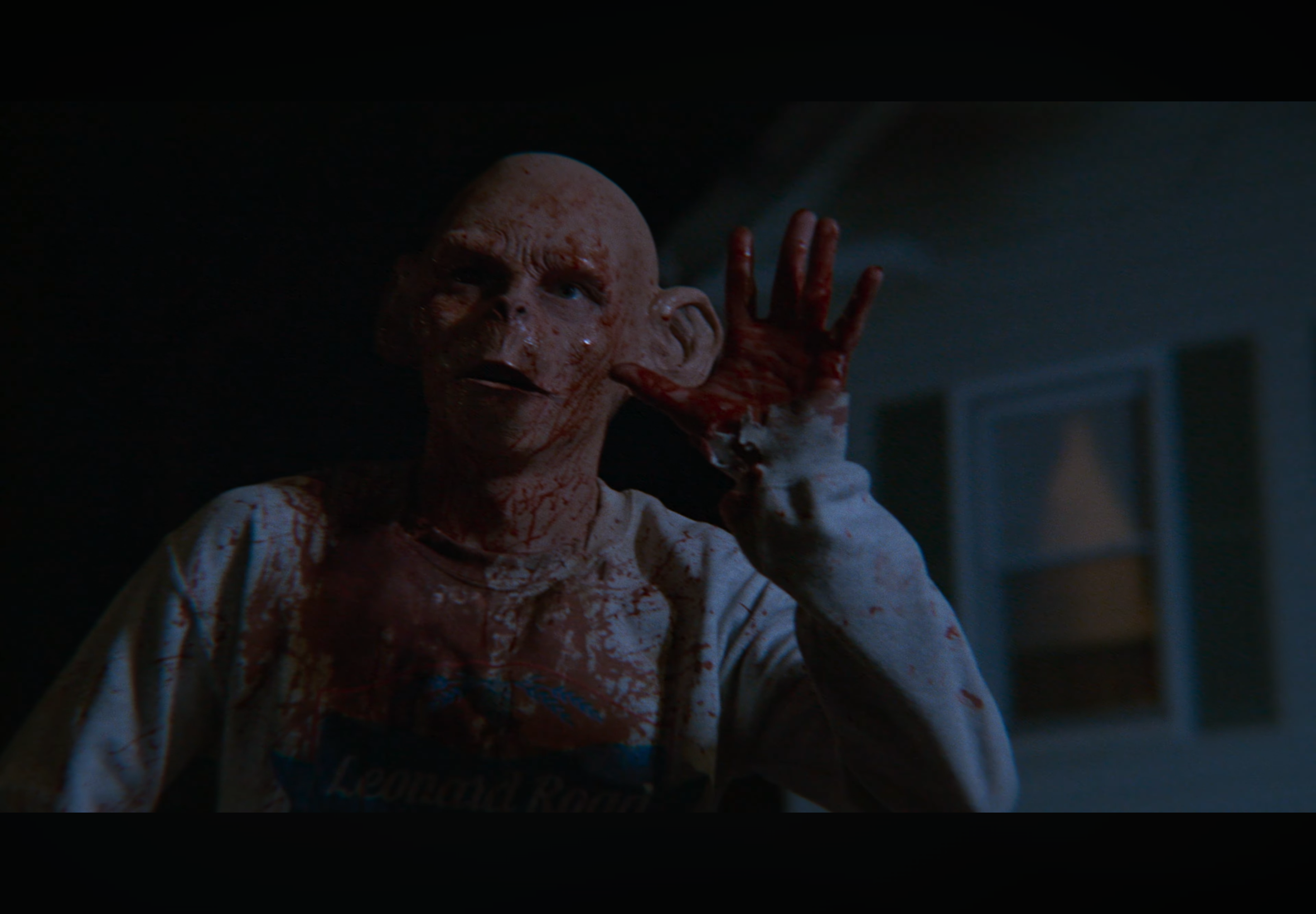 A person with a bald head and blood on their face and shirt makes a telephone gesture with their hand.