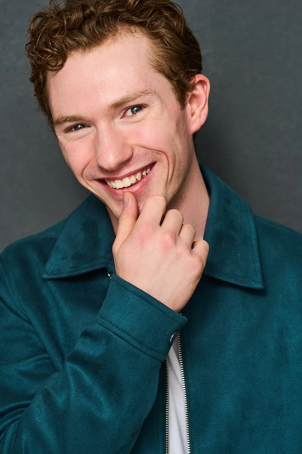 A young man with red curly hair and blue eyes smiling and touching his chin with his right hand, wearing a teal jacket against a gray background.