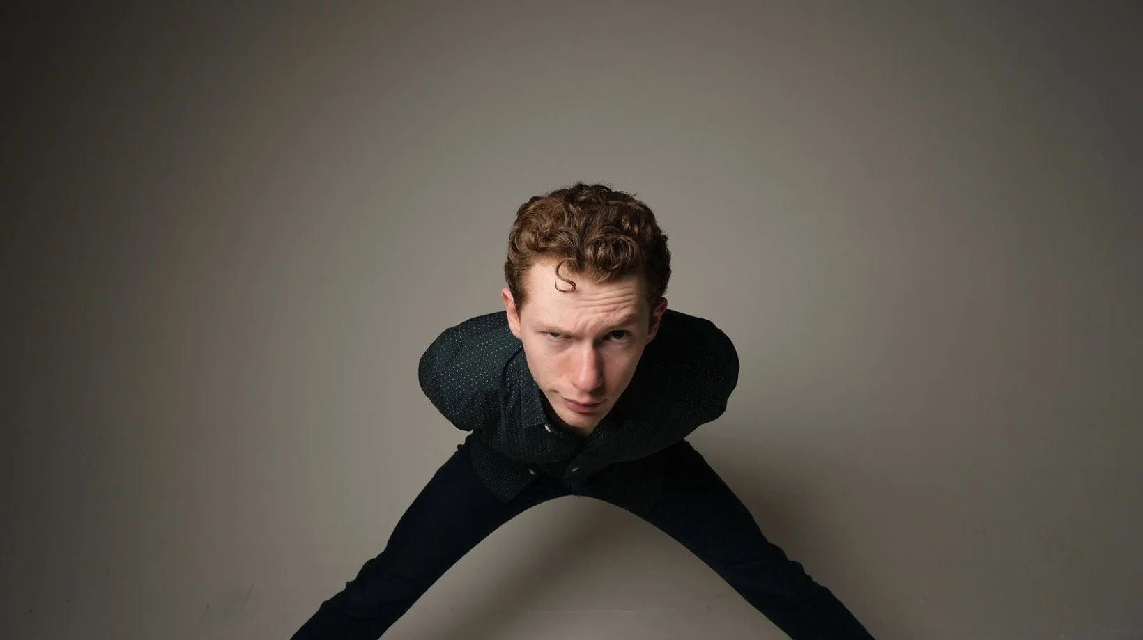 A young man with curly hair in black clothing squats with his legs apart and leans forward, looking directly at the camera with a serious expression against a plain, gradient background.