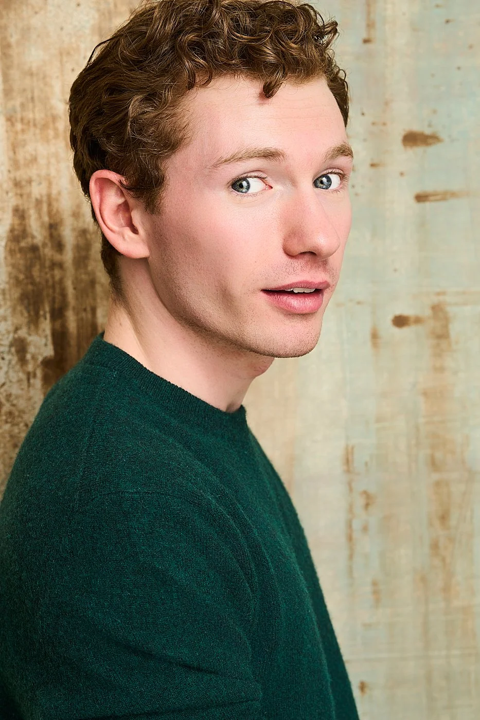 A young man with light skin, curly light brown hair, and blue eyes, wearing a dark green sweater, posing in front of a rustic wooden background.