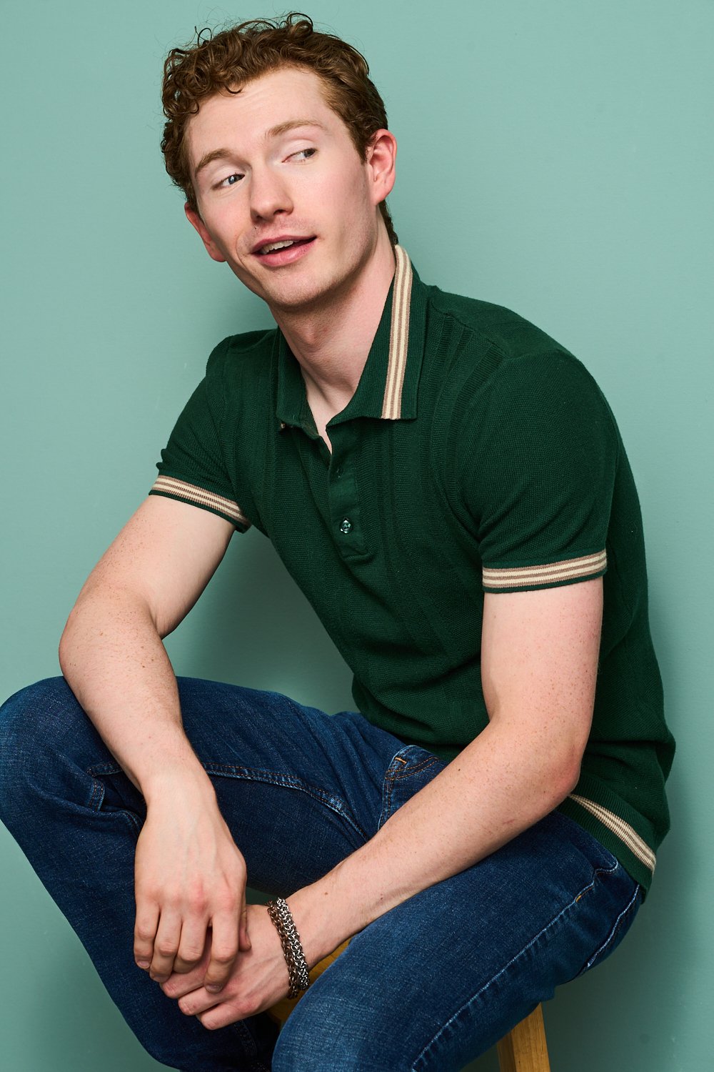 A young man with curly red hair and fair skin is sitting on a stool against a plain light green background. He is wearing a dark green polo shirt with beige and brown striped trim, blue jeans, and a silver chain bracelet on his wrist. He has a friendly expression, with his head turned slightly to the side and a slight smile.