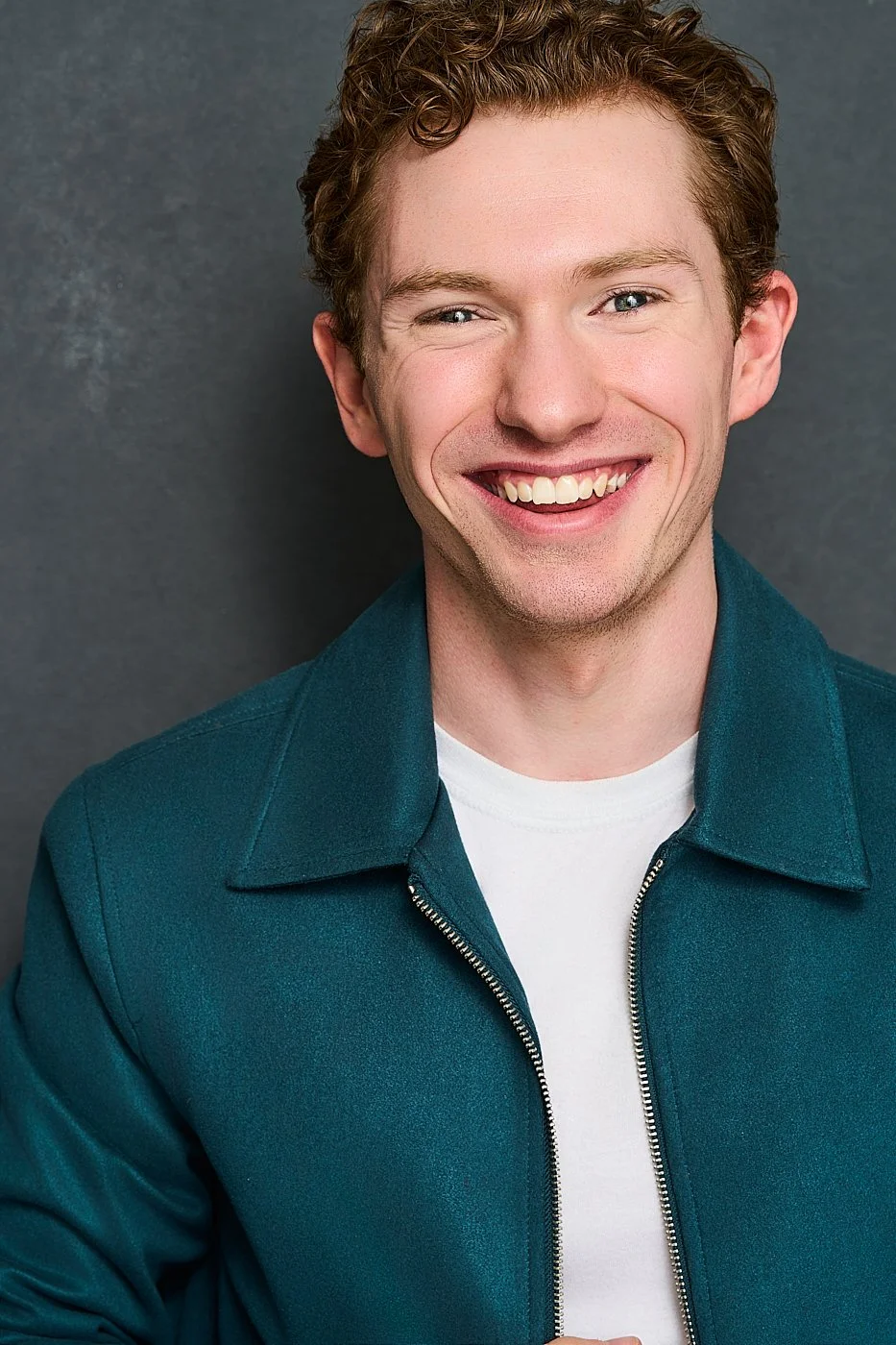 A young man with curly red hair and fair skin smiling brightly, wearing a teal zip-up jacket over a white shirt, standing against a gray background.