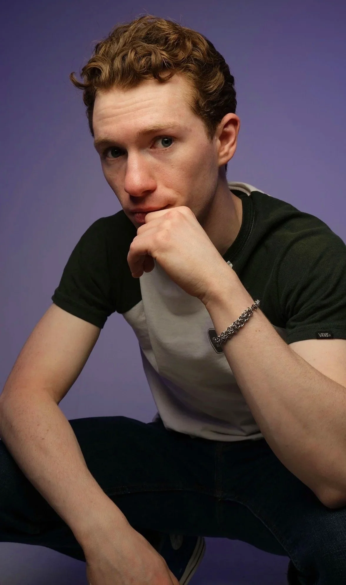 A young man with curly red hair and light skin is sitting against a purple background, resting his chin on his hand, wearing a black and white T-shirt with a small logo on the sleeve and a silver chain bracelet on his wrist.