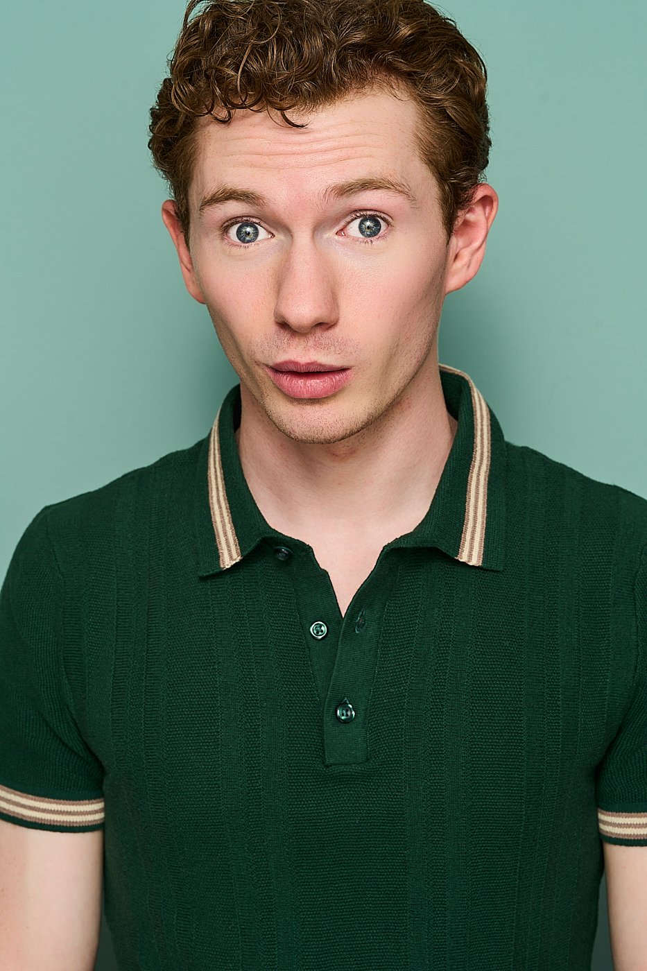 A young man with curly light brown hair and blue eyes wearing a green polo shirt with striped collar and sleeve cuffs posing in front of a light green background.