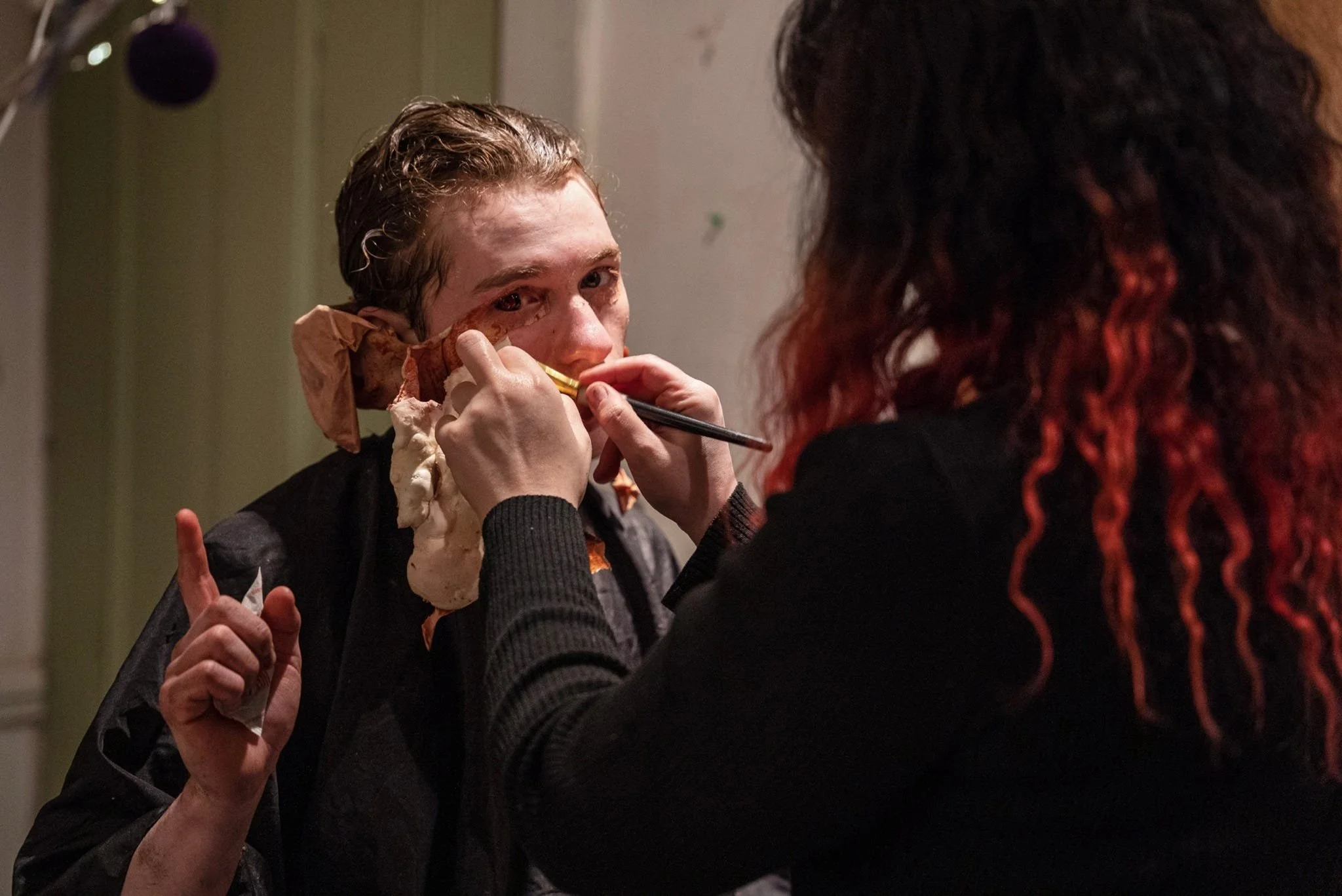 A person with red hair applies makeup to Zach Norton in a dimly lit room.
