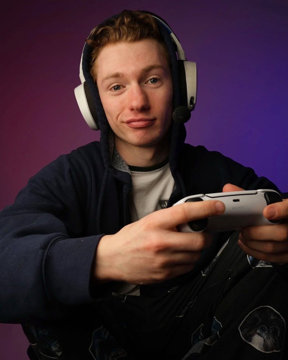 Zach Norton wearing a gaming headset and hoodie, holding a game controller, smiling with purple background.