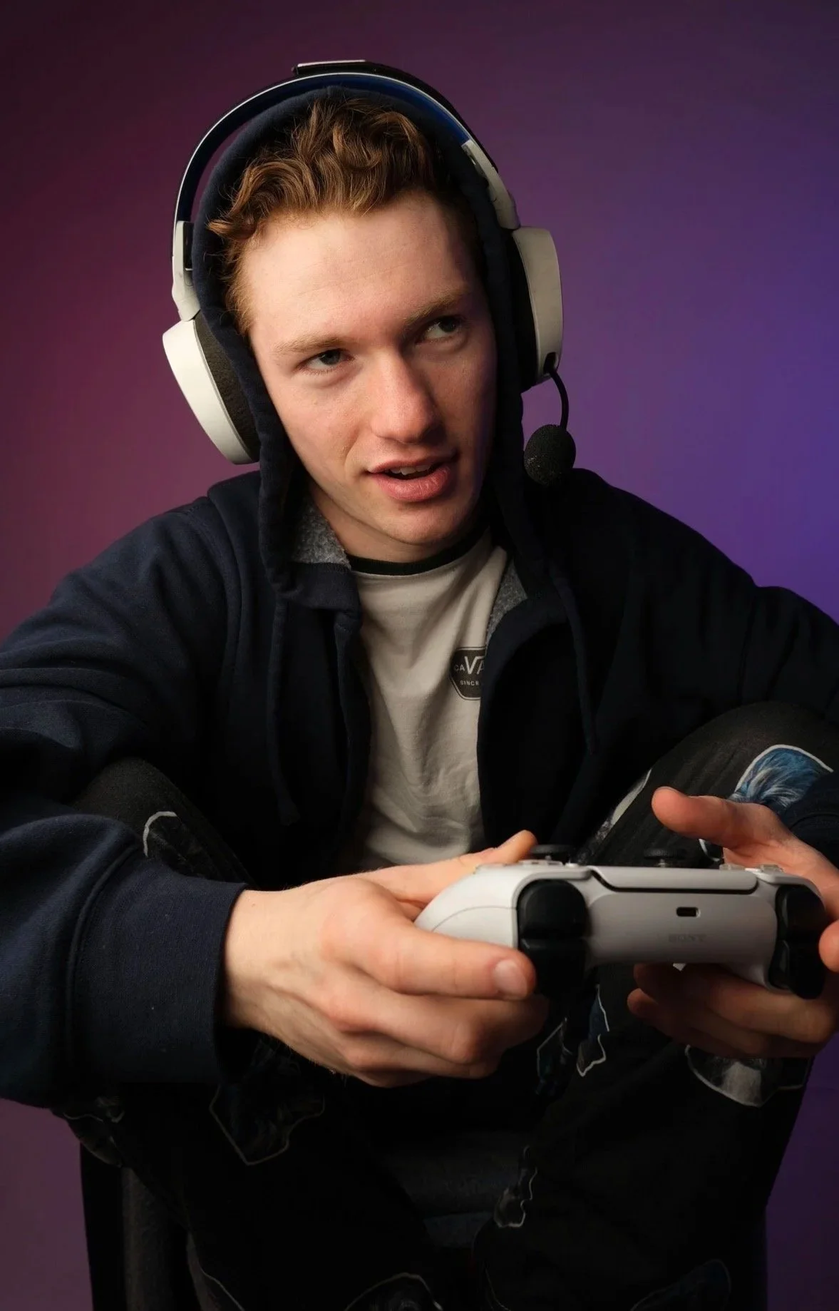 A young man with red hair wearing a gaming headset with a microphone, holding a video game controller, sitting against a purple background.