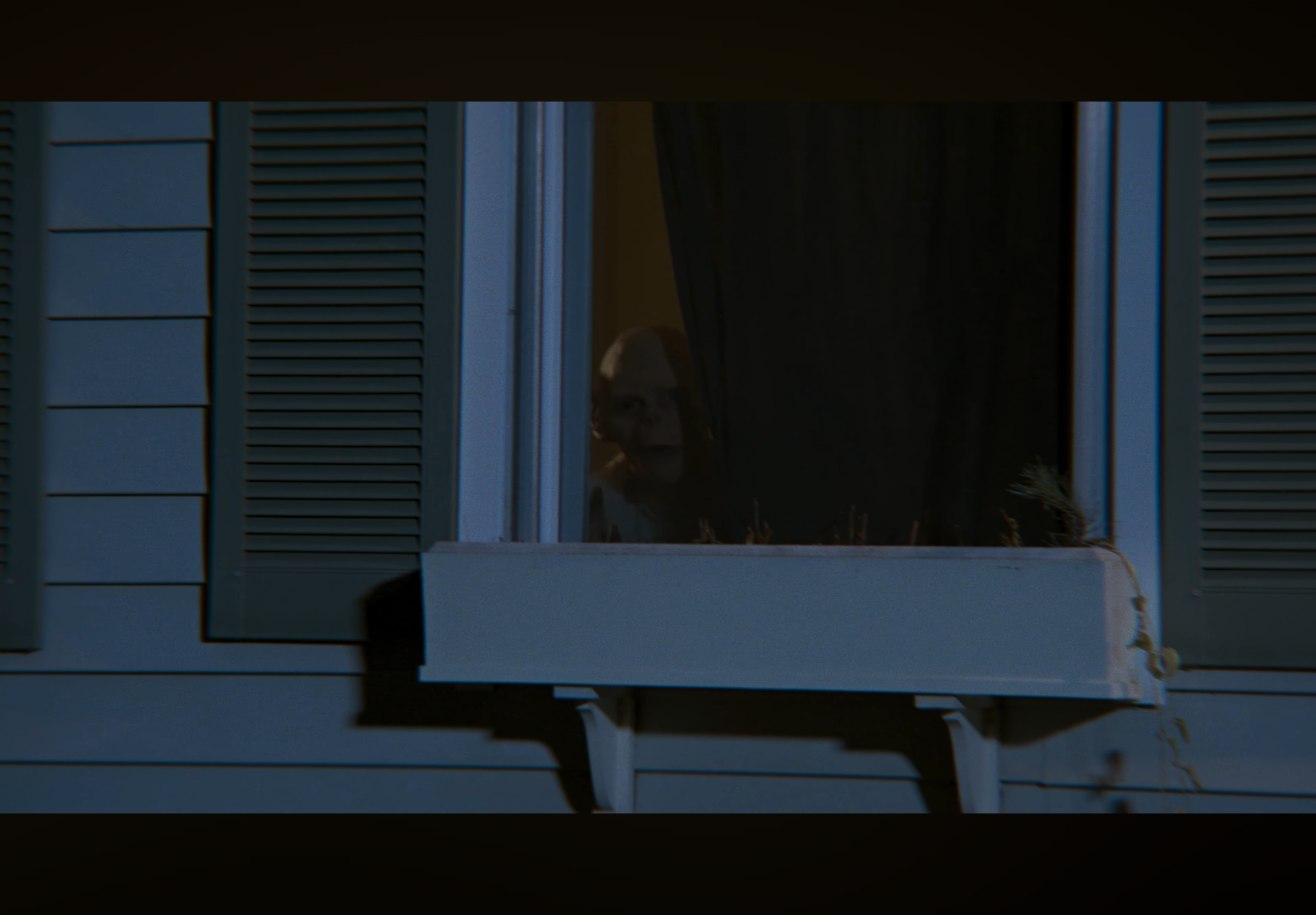 A person looking out from a window at night, dark indoors, with the window shutters partially open.