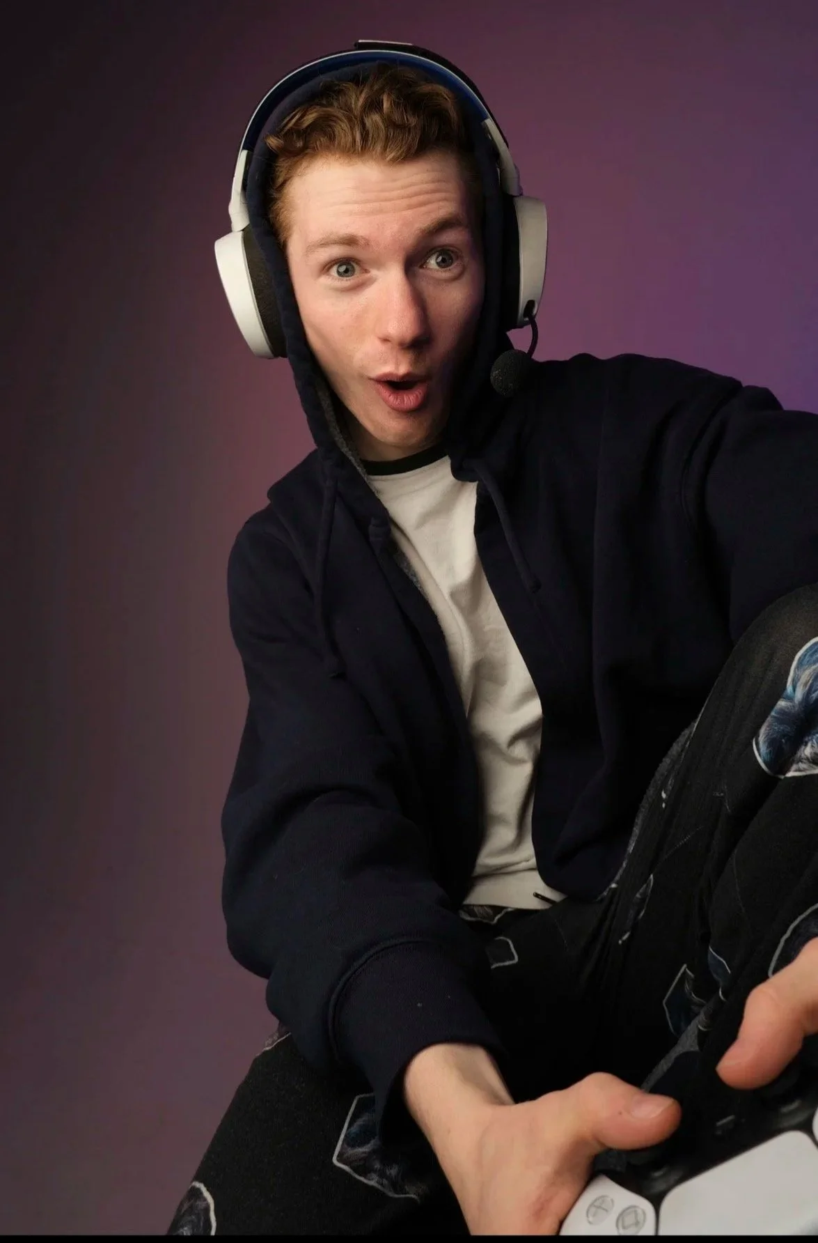 A young man with curly reddish hair wearing headphones with a microphone, making a surprised face while holding a gaming controller.