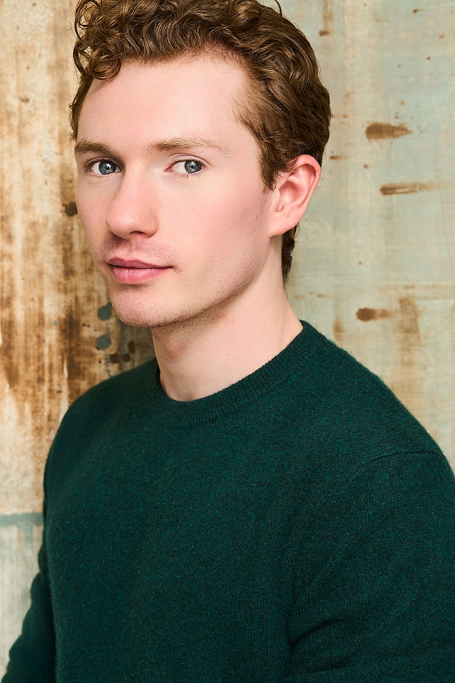A young man with curly brown hair and blue eyes, wearing a dark green sweater, posing against a rustic wooden background.