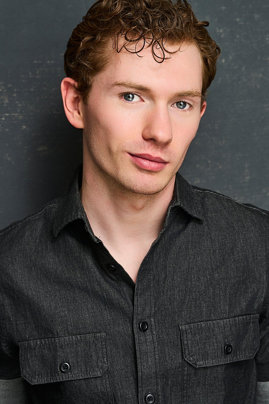 Portrait of a young man with curly brown hair and blue eyes, wearing a dark denim shirt, standing against a dark textured background.