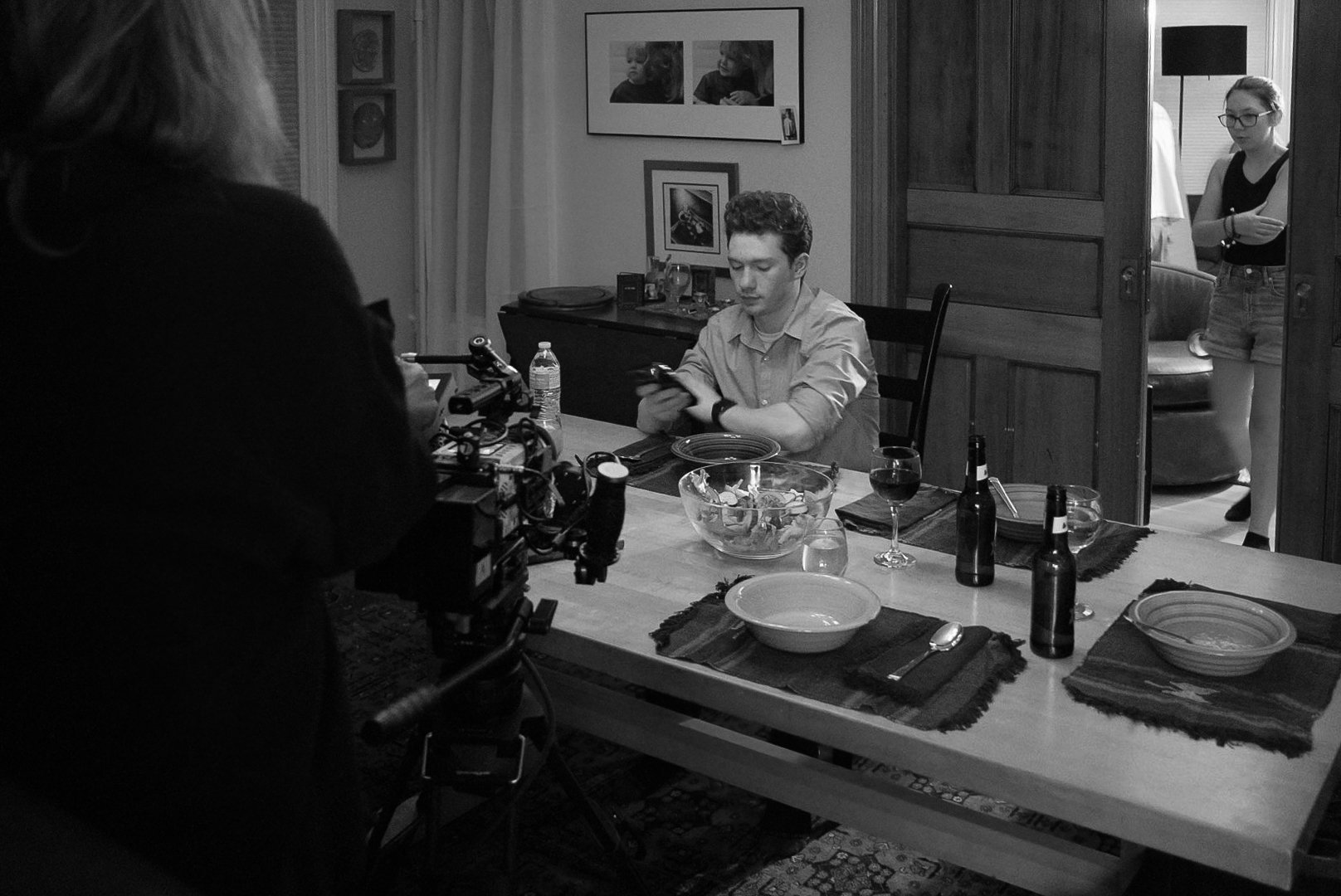 A man sitting at a dining table, looking at his phone, with drinks and bowls in front of him. Behind a partially open door, a woman stands in the other room, and a woman in glasses looks at her phone.