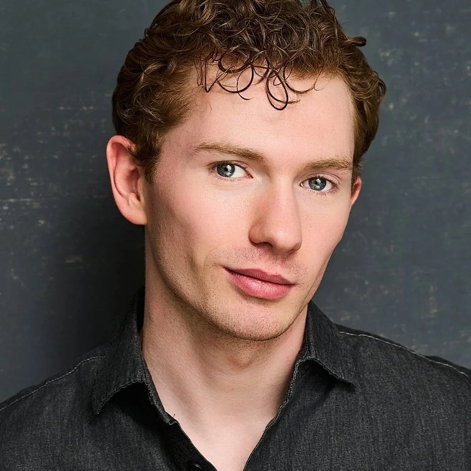 Close-up portrait of Zach Norton with curly light brown hair and blue eyes, wearing a dark shirt, standing against a dark textured background.