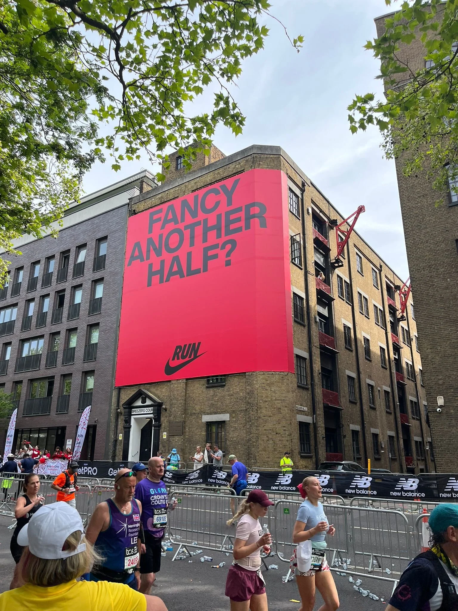 A group of marathon runners participating in a race on a city street, with a pink billboard advertising Nike in the background reading 'FANCY ANOTHER HALF?'. The street is lined with barriers, and there are trees and buildings overhead.