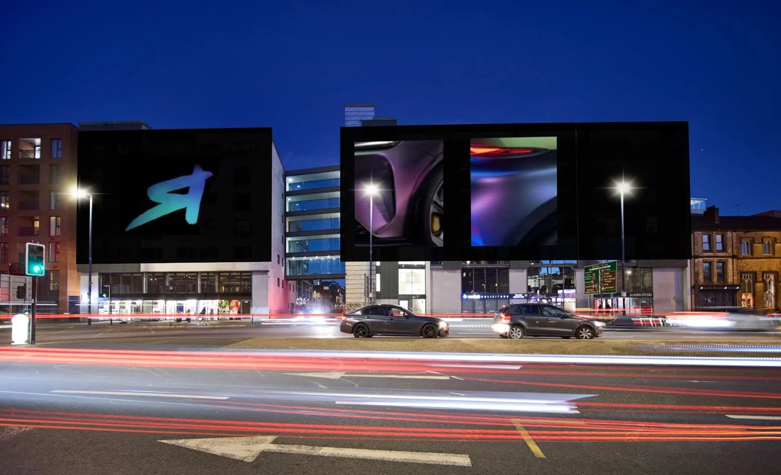 Nighttime city street view with moving cars creating light trails, modern building with digital billboards displaying logos and images, and traffic signals.