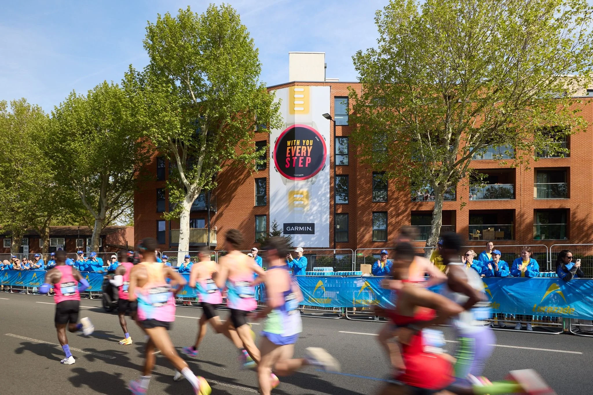 Runners participating in a marathon, with some wearing pink shirts and purple socks, passing by spectators and a large advertisement display on a red-brick building. The scene takes place on a city street with green trees and a clear blue sky.