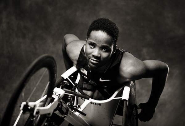 A male athlete in a wheelchair, looking up at the camera with a determined expression.