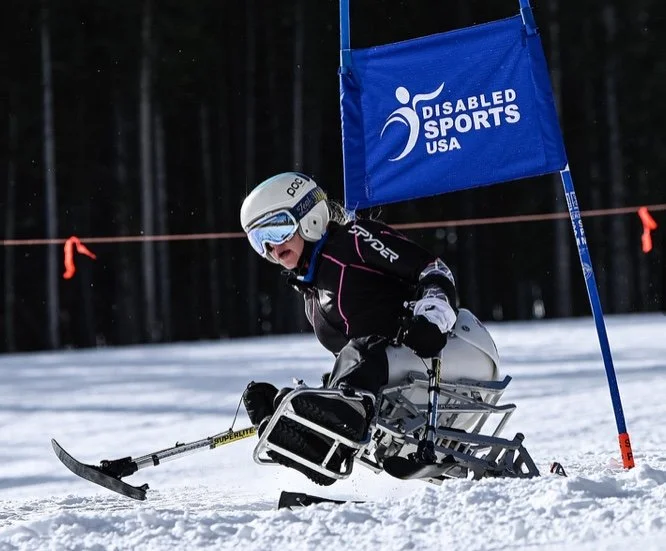 A skier in a racing wheelchair navigating a snowy course marked by a blue flag with the Disabled Sports USA logo.