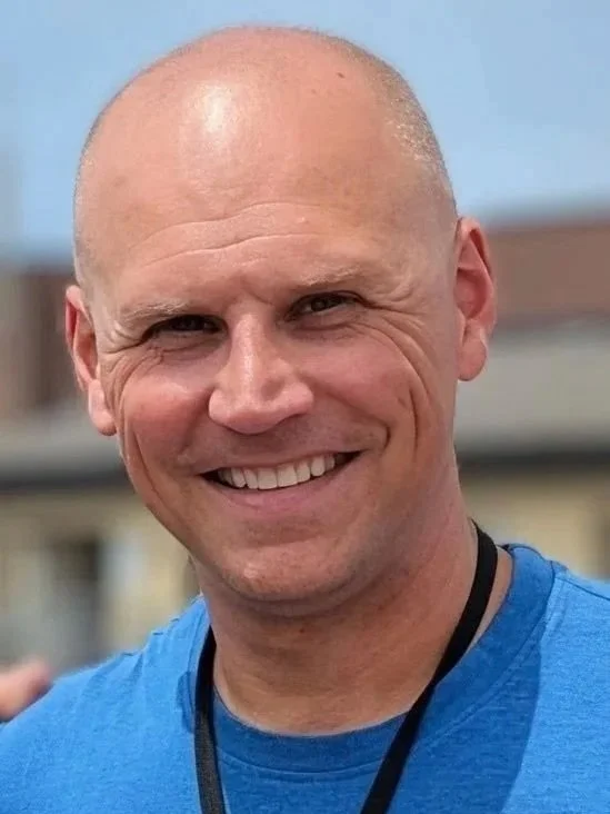 A smiling man with a shaved head wearing a blue shirt and a lanyard, outdoors on a cloudy day.