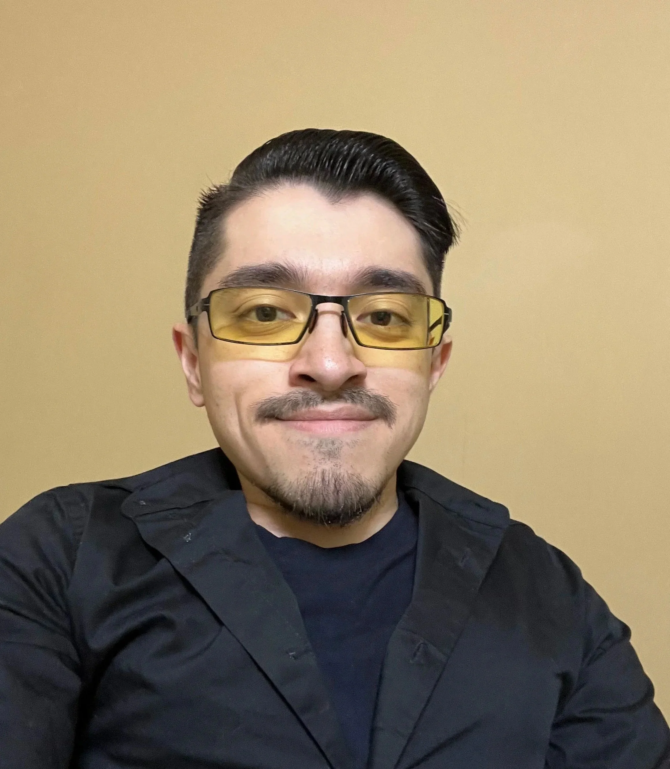 Close-up of a man wearing yellow-tinted glasses, a mustache and goatee, with dark, short hair styled upward, smiling slightly, against a beige wall background, dressed in a black jacket.