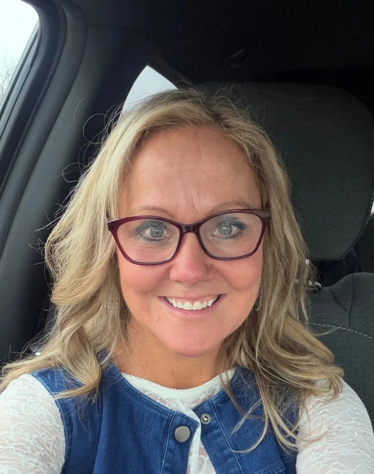 A woman with blonde hair, glasses, and a bright smile, sitting in a car seat.