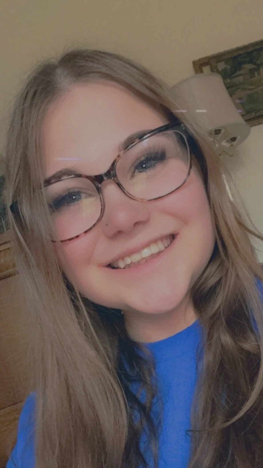 A smiling young woman with long, light brown hair and glasses, wearing a blue shirt, in a room with beige walls and a lamp behind her.