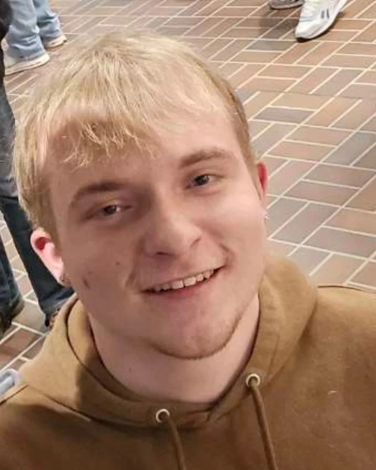 A young man with light brown hair smiling, wearing a brown hoodie, standing on a tiled floor with other people around.