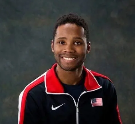 Portrait of a smiling Black man with short curly hair, wearing a black athletic jacket with red and white accents, featuring an American flag patch and Nike logo, against a dark, textured background.