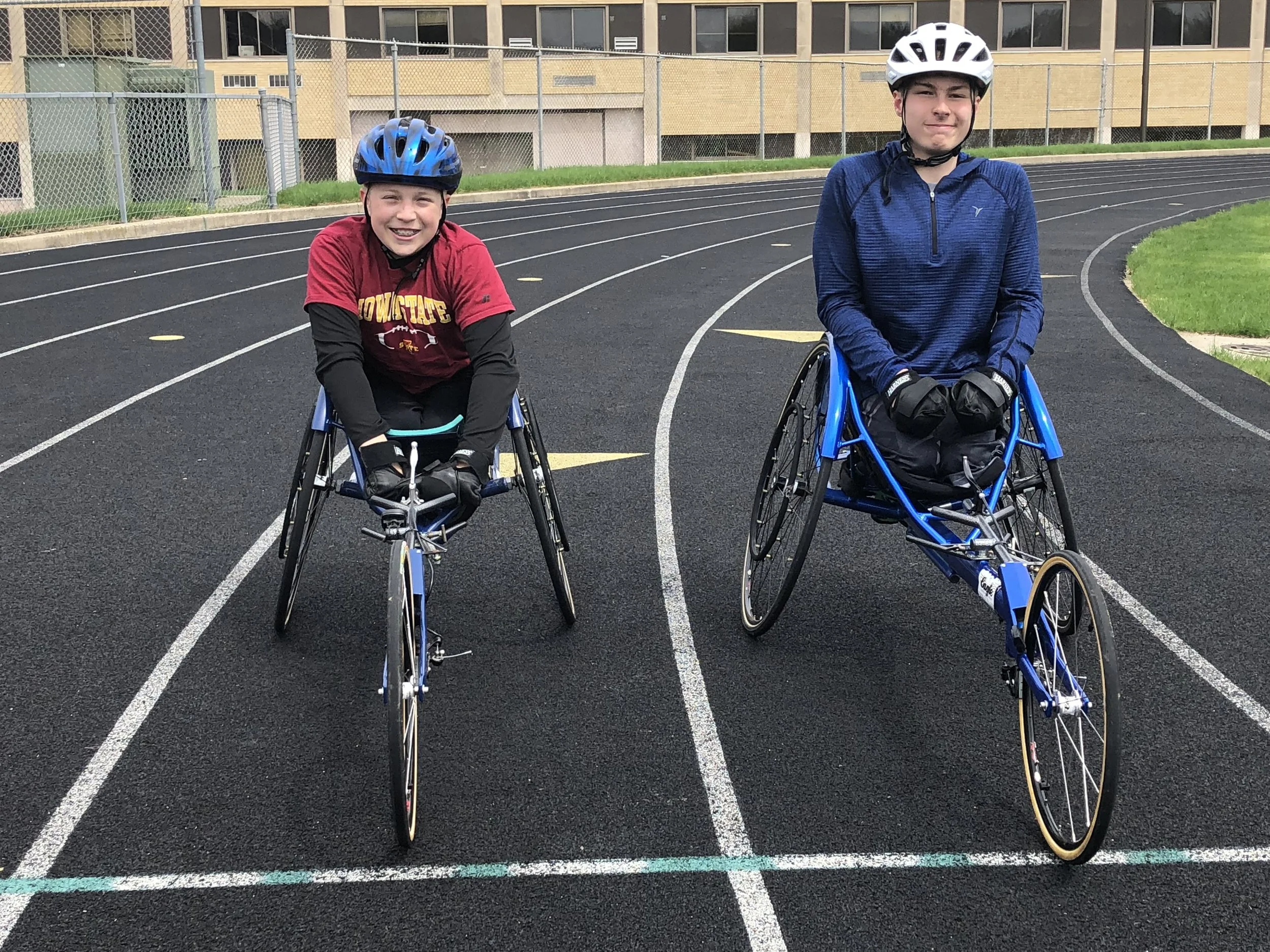 Two young boys in wheelchairs on a running track. The boy on the left wears a red shirt, black long sleeves, and a blue helmet, smiling at the camera. The boy on the right wears a blue jacket and a white helmet, looking at the camera with a slight smile. There is a yellow building and a chain-link fence in the background.