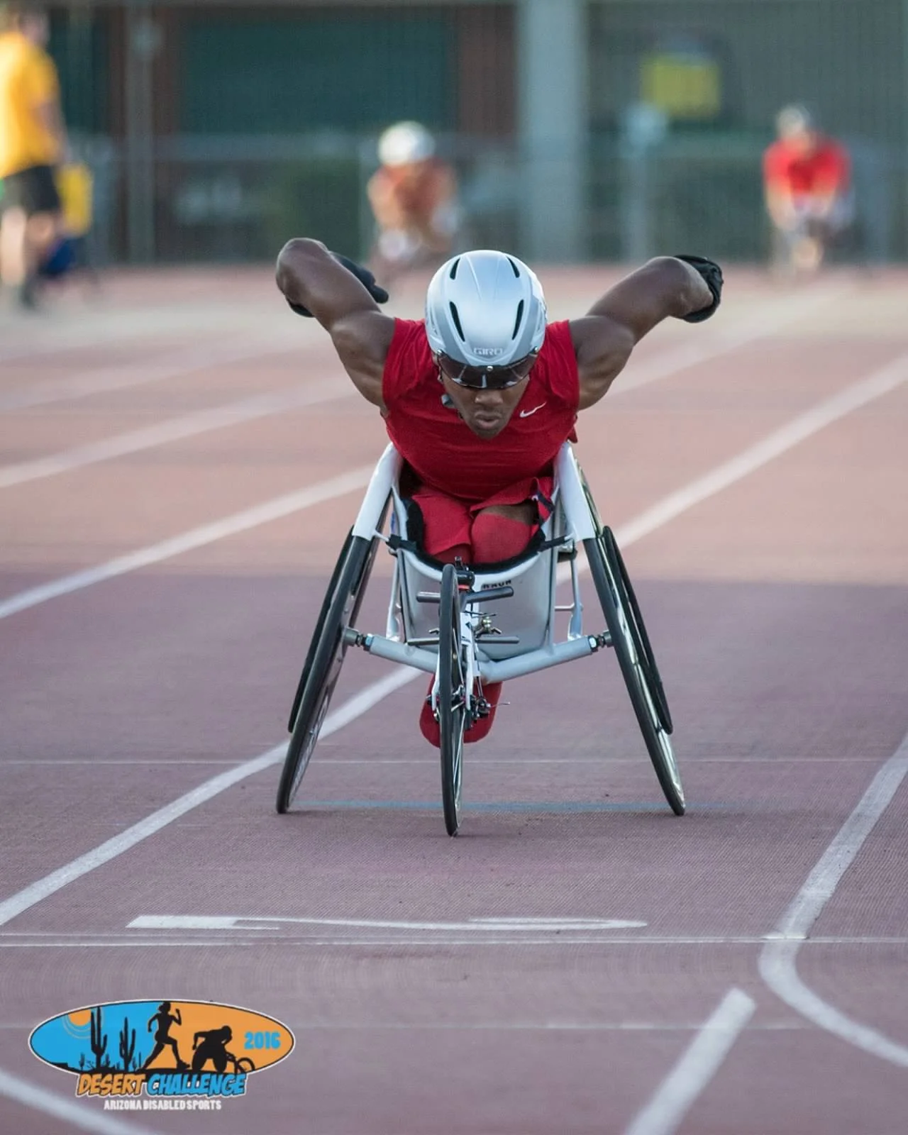 A male athlete in a wheelchair preparing to race on a track, wearing a helmet, sunglasses, and a red athletic outfit, with other athletes in the background. There is a logo on the track indicating the 2016 Desert Challenge for Arizona Disabled Sports
