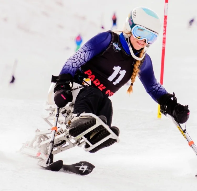 A female adaptive skier in a wheelchair glides on snow, wearing a helmet, goggles, and a blue and black suit, participating in a ski race.