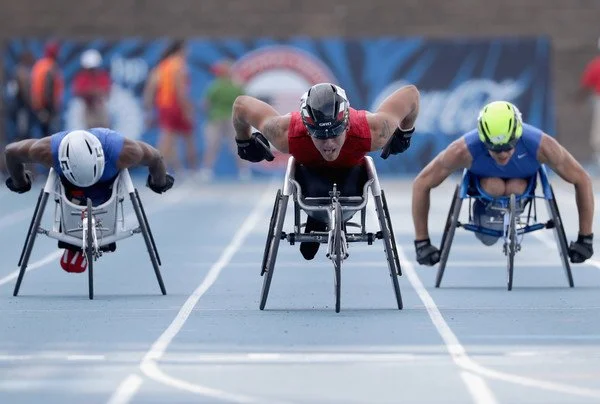 Three wheelchair racers in a sprint race on a blue track, wearing helmets and athletic gear, pushing their wheelchairs forward.