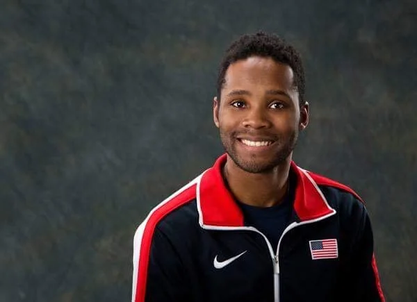 Portrait of a smiling man wearing a red, black, and white Nike sports jacket with an American flag patch, against a dark mottled background.