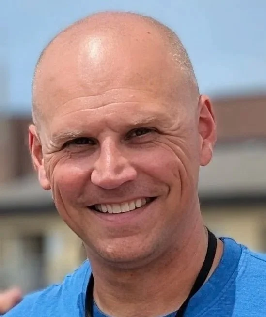 A smiling bald man wearing a blue shirt with a blurred outdoor background.