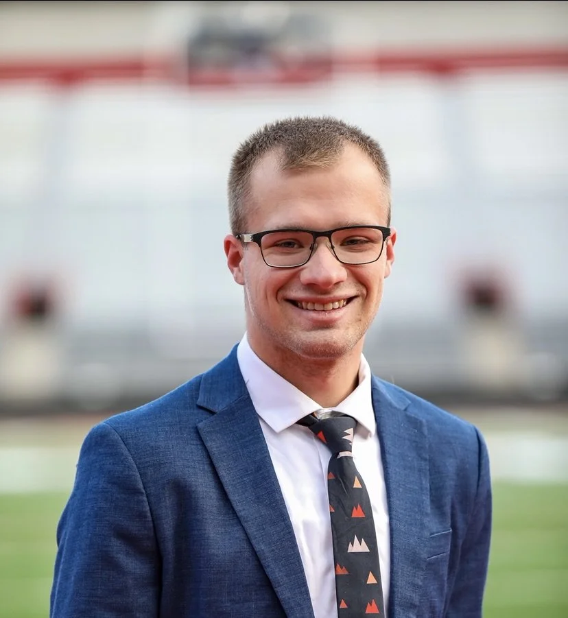 A young man in a blue suit, white shirt, and patterned tie smiling outdoors with blurred stadium background.