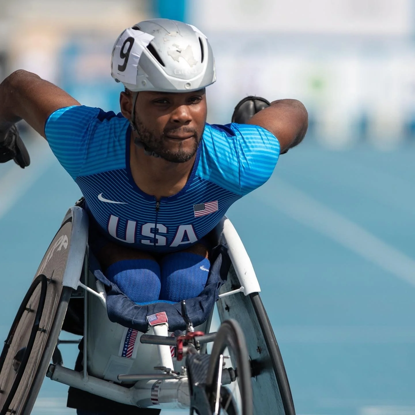 A male athlete in a wheelchair wearing a blue USA sports uniform and helmet, preparing for a race on a track.