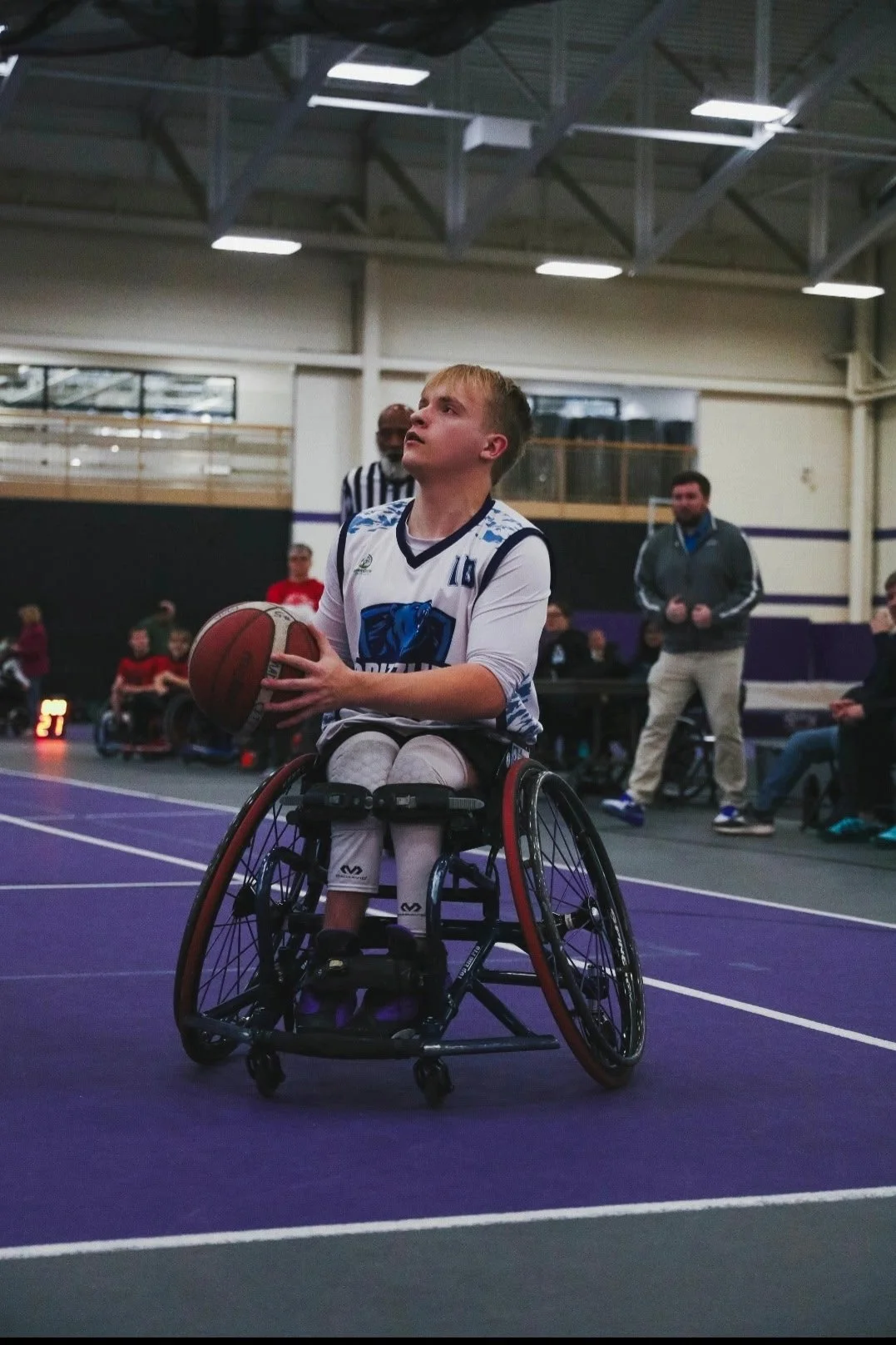 A young person in a wheelchair playing basketball indoors on a purple court, holding a basketball, with a focused expression. People and coaches are visible in the background.
