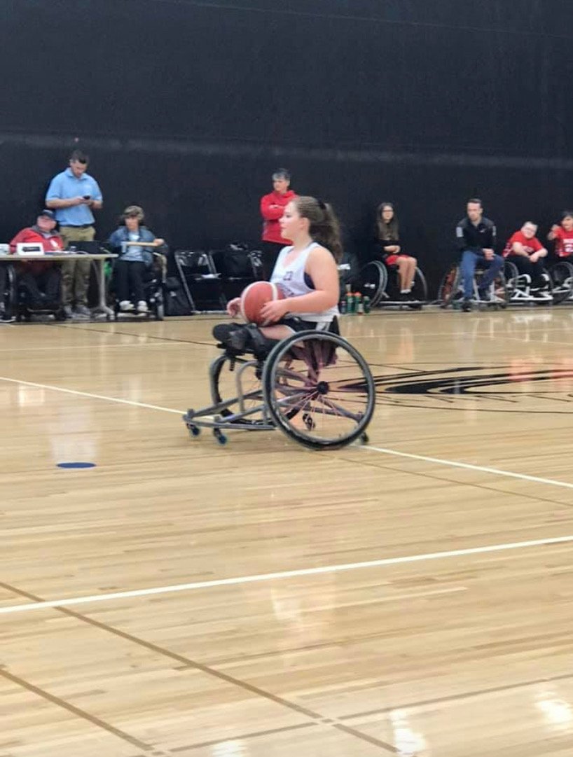 A female athlete in a wheelchair holding a basketball on a gymnasium basketball court, with spectators and officials seated in the background.