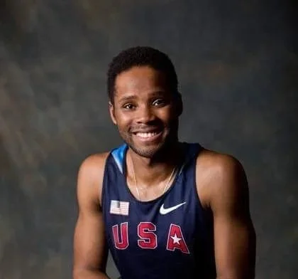 A smiling male athlete wearing a USA athletic jersey with the American flag patch, set against a dark background.
