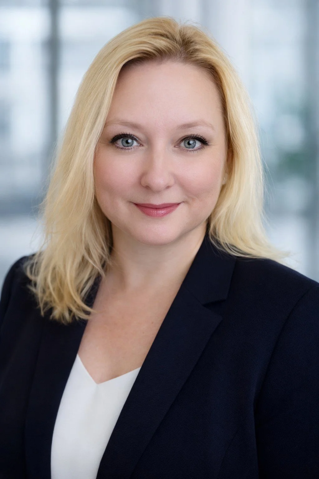 Professional headshot of a smiling blonde woman with blue eyes, wearing a black blazer and a white top, in an office setting.