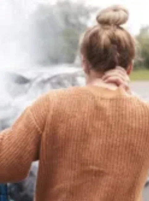 Back view of woman with her hair in a bun, wearing a brown sweater, touching her neck, outside near a car.